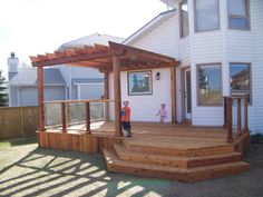 two children are standing on the front porch of a house that has a wooden deck and pergolated area