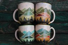 four coffee mugs with mountains and trees painted on them sitting on a wooden table