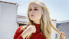 a woman with long blonde hair standing in front of a white building wearing a red top