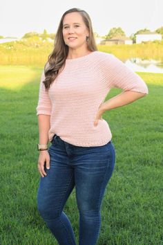 a woman standing in the grass with her hands on her hips, wearing jeans and a pink sweater