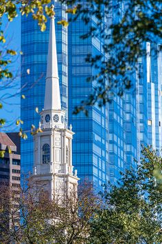 a church steeple in front of tall buildings with trees and blue sky behind it