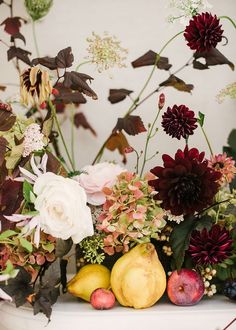 an arrangement of flowers, fruit and leaves is displayed on a shelf in front of a wall