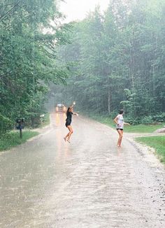 two people running in the rain on a road with trees behind them and one person holding an umbrella