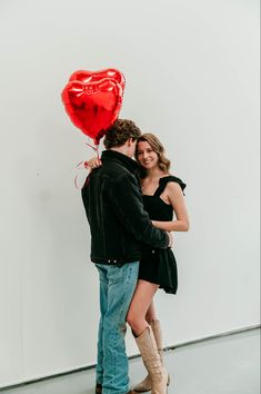 a man and woman standing next to each other in front of a white wall with a red heart balloon