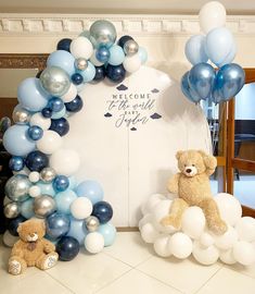 two teddy bears sitting in front of a welcome to the world sign and balloon arch