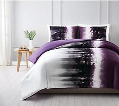 a bed with purple and white comforters on top of it, next to a plant