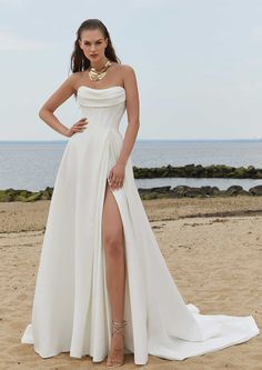 a woman standing on top of a sandy beach wearing a white dress with high slit