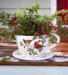 a cup filled with holly and birds sitting on top of a wooden table covered in snow