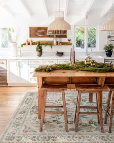 a wooden table topped with lots of greenery next to a kitchen island covered in hanging lights