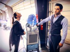 a man and woman standing next to an air conditioner in a garage with the door open