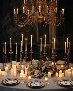 a table topped with lots of candles and plates covered in white flowers next to a chandelier