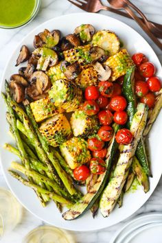 a white plate topped with asparagus, tomatoes and grilled vegetables next to glasses of green juice