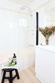 a bathroom with white walls and tile flooring has a vase on the stool next to the shower