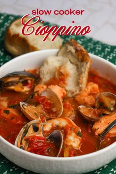 a white bowl filled with seafood and clams in tomato sauce next to bread on a green towel