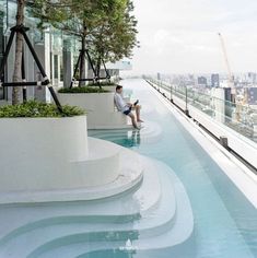 a man sitting on the edge of a swimming pool next to tall buildings and trees