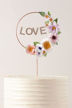 a white cake with flowers on top and the word love spelled in gold letters above it