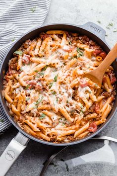 a skillet filled with pasta and sauce on top of a towel next to a wooden spoon