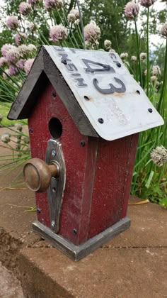 a red birdhouse sitting on top of a cement slab next to flowers and grass