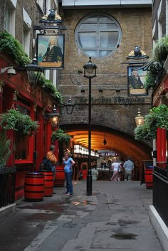 people are walking down an alley way with red barrels on either side and hanging plants