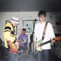 two young men are playing guitars and singing into microphones in a room with other people