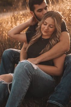 a man and woman sitting on the ground in front of some tall grass with their arms around each other