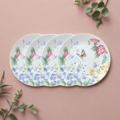 four plates with flowers and butterflies on them sitting next to green leaves in front of a pink wall