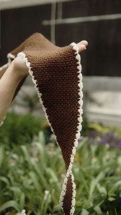 a hand holding up a brown and white knitted hat in front of some flowers