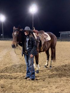 a woman standing next to a brown horse on top of a dirt field at night