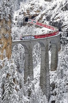 a red train traveling over a bridge in the middle of snow covered mountains and trees