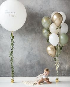 a baby sitting on the floor in front of balloons and greenery, while another balloon is floating above it