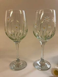 two wine glasses sitting next to each other on top of a white tablecloth with daisies painted on them