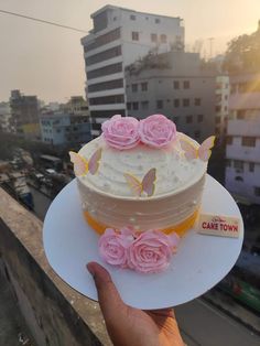 a hand holding a cake with pink roses on it in front of a cityscape