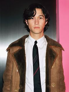 a young man wearing a brown jacket and tie standing in front of a pink wall