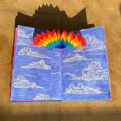 an open book with a rainbow and clouds drawn on the pages, sitting on a sidewalk