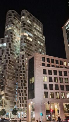 some very tall buildings at night time with lights on and people crossing the street in front