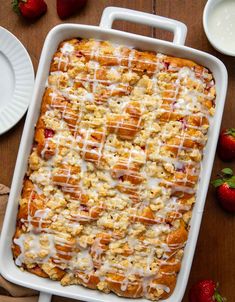 a white dish filled with bread and covered in icing next to strawberries on a wooden table