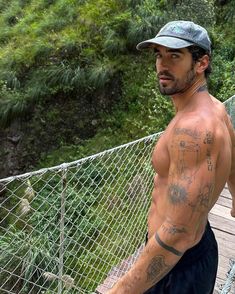 a shirtless man standing on a bridge with trees in the background and bushes behind him