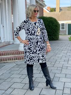 an older woman standing on a brick walkway wearing black boots and a dress with white swirls