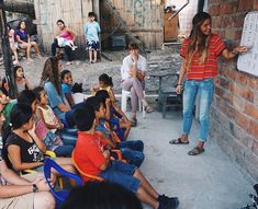 a woman standing in front of a group of children sitting on chairs and writing on a whiteboard