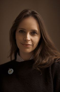 a woman with long brown hair and a black sweater smiles at the camera while wearing a badge on her collar