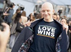 a bald man in a love wins t - shirt is holding up his jacket and smiling at the camera