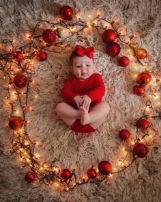 a baby in a red bodysuit sitting on a rug surrounded by christmas lights