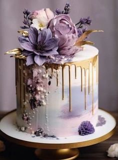 a white cake with purple flowers and gold drips on the top is sitting on a table