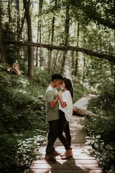 a man and woman standing on a wooden walkway in the woods with their arms around each other