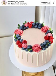 a cake decorated with berries and blueberries on top of a white table next to a window