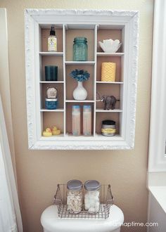 a white toilet sitting next to a bathroom sink under a mirror above it is a shelf filled with jars and containers