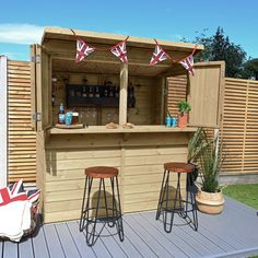 an outdoor bar with two stools and some flags on the back wall, next to a wooden fence