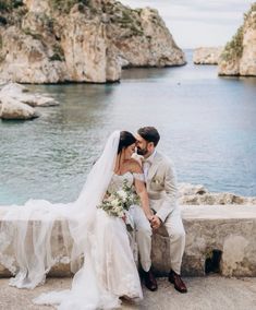 a bride and groom sitting on a stone wall by the water