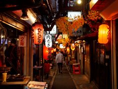 a man walking down an alley way with lanterns hanging from it's ceiling and lights on either side