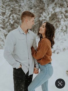 a man and woman standing in the snow with trees behind them, looking at each other